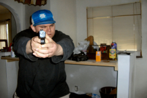 Ein Mann hält eine Waffe mit beiden Händen und steht vor einem Tisch mit verschiedenen Gegenständen, mit einer Wand und zwei Fenstern im Hintergrund.