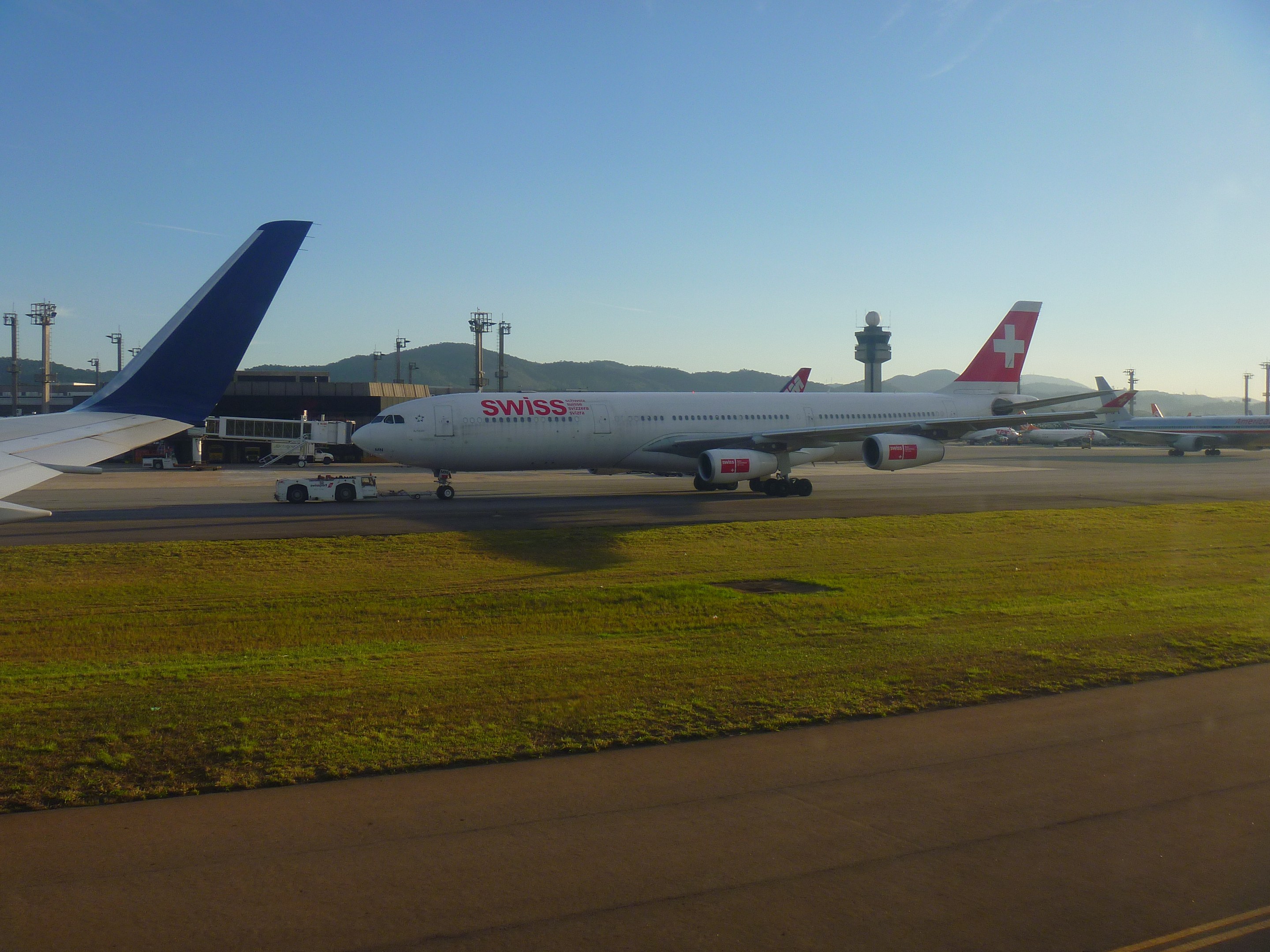 Schweizer International Air Lines Boeing 777-300ER auf der Rollbahn am San Francisco International Airport, mit Gras, einer Straße, Laternenmasten, Gebäuden, Hügeln und einem klaren blauen Himmel im Hintergrund.