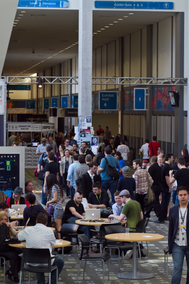Ein belebter Flughafen-Vorhalle mit Wartenden, einige stehen und andere arbeiten, Richtungsschilder an der rechten Wand und darüber, sowie von der Decke hängende Lampen.