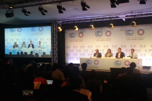 Menschen sitzen auf Stühlen auf einer Bühne mit Namensschildern und Mikrofonen auf einem Tisch davor, während Zuschauer von Stühlen aus zusehen; ein Projektionsschirm und Beleuchtung an Stangen sind im Hintergrund sichtbar.