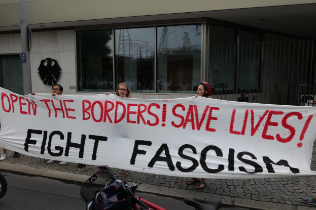 A group of people holding a banner reading "Open the Borders, Save Lives, Fight Fascism" in front of a building with glass windows and a pole, with a bicycle and bag on the road.