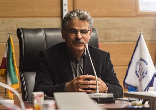 A man in a black blazer and glasses sits at a table with a microphone, surrounded by multiple flags, in front of a wooden wall with a window; he is identified as the Minister of the Interior of the Ministry of Foreign Affairs.