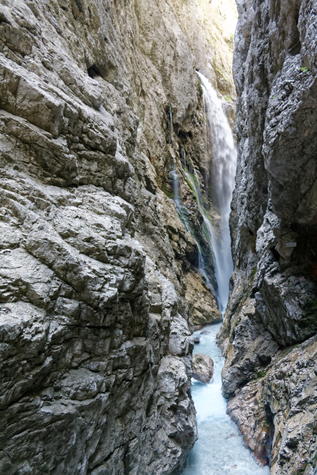 Kleiner Wasserfall, der über zerklüftete Felsen in einem steinigen Tal hinabfließt, umgeben von saftig grünen Hügeln unter strahlendem Sonnenschein.