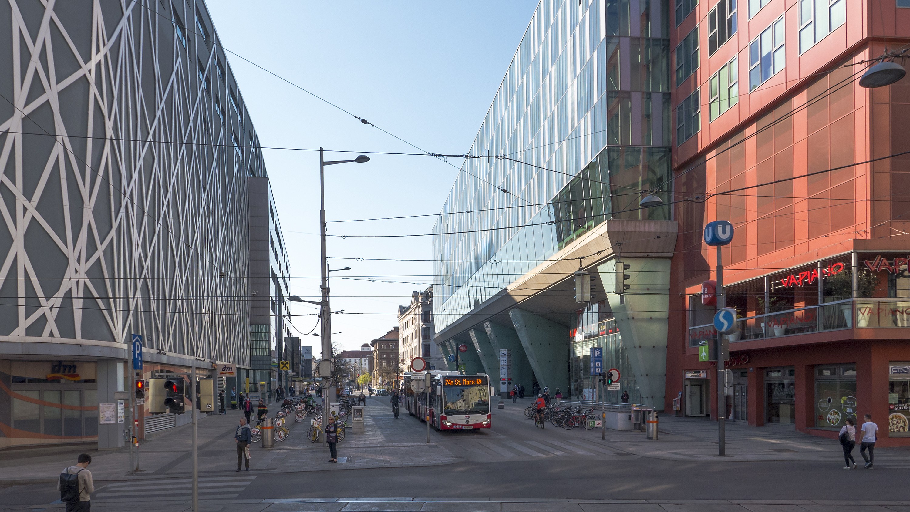 A city street with tall buildings, street poles, lights, electric cables, vehicles, pedestrians, bicycles, signs, and a clear blue sky.