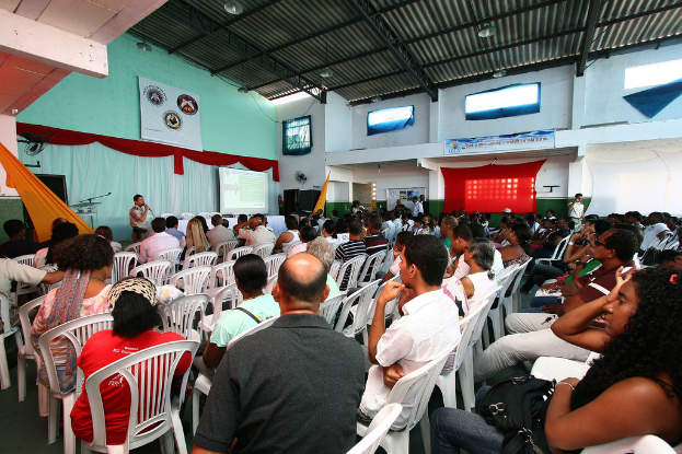 Eine Gruppe von Menschen, die auf Stühlen sitzen und einer Buhne gegen√∫berblicken, auf der ein Polizeibeamter in ein Mikrofon spricht, mit einer T√ľr auf der rechten Seite des Bildes.