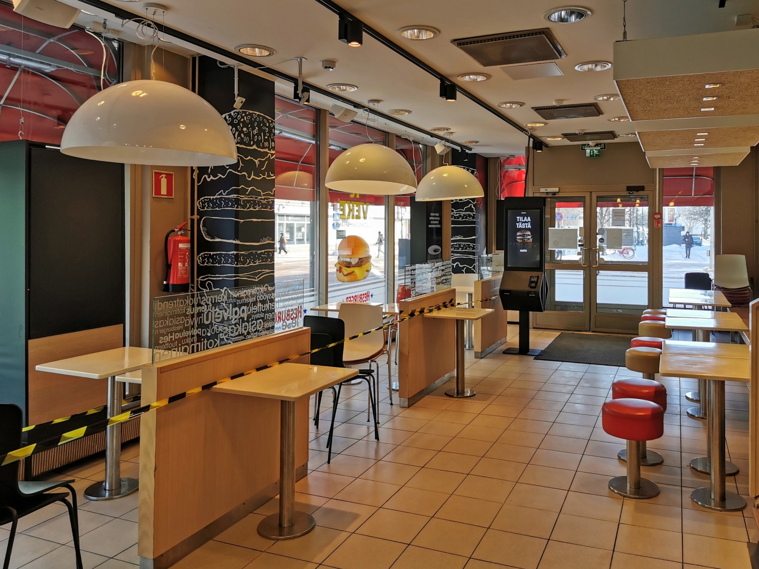 Interior of a fast food restaurant with tables, chairs, stools, a floor, ceiling lights, a wall-mounted fire extinguisher, glass doors, and additional furnishings.