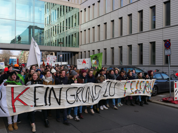 Eine Gruppe von Menschen steht auf einer Straße und hält ein Transparent mit der Aufschrift "Kein Grind Lo Feuer" hoch, umgeben von Gebäuden, Bäumen, Pfählen, Schildern, Fahrzeugen und einem klaren blauen Himmel.