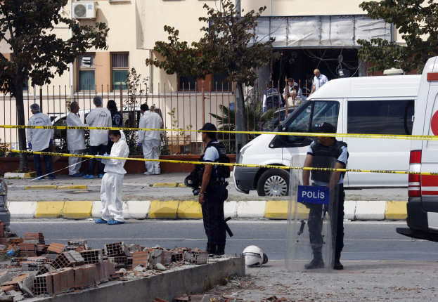 Eine Gruppe von Polizeibeamten steht vor einem Gebäude mit Fenstern, umgeben von Fahrzeugen, einem Zaun, Bäumen und Pflanzen, mit Ziegelsteinen und einem Helm im Vordergrund, wahrscheinlich nach einer Bombenexplosion in Mexiko-Stadt.