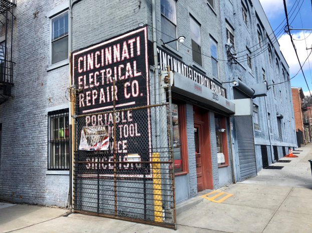 Außenansicht des Cincinnati Electrical Repair Co. Gebäudes mit Beschriftung, einem Metallzaun, einem Müllbehälter, Versorgungsmasten und -drähten, Bäumen und einem bewölkten Himmel.
