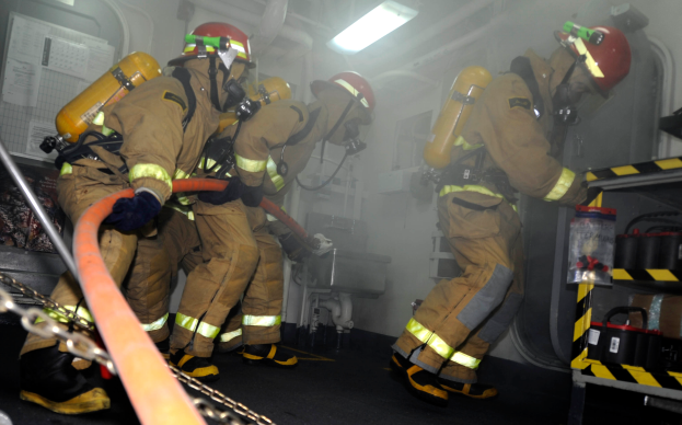 Eine Gruppe von Feuerwehrleuten in Schutzausrüstung, darunter Helme und Sauerstoffflaschen, arbeitet an einem Feuerwehrauto. Rechts davon befindet sich ein Gestell mit verschiedenen Gegenständen und im Hintergrund Rohre, Lichter und andere Gegenstände.