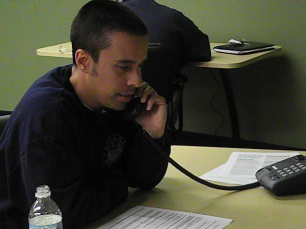 Mehrere Personen sitzen um einen Tisch, wobei eine Person auf einem auf dem Tisch liegenden Telefon spricht, zusammen mit einem stationären Gerät, einer Einwegflasche und Kabeln.