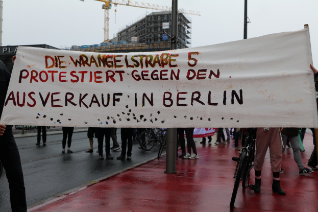 A group of people holding a banner that reads "Die Wrangellstraße 5 Protestiert Gegen den Ausverkauf in Berlin" on a road with bicycles, poles, buildings, and a crane in the background.