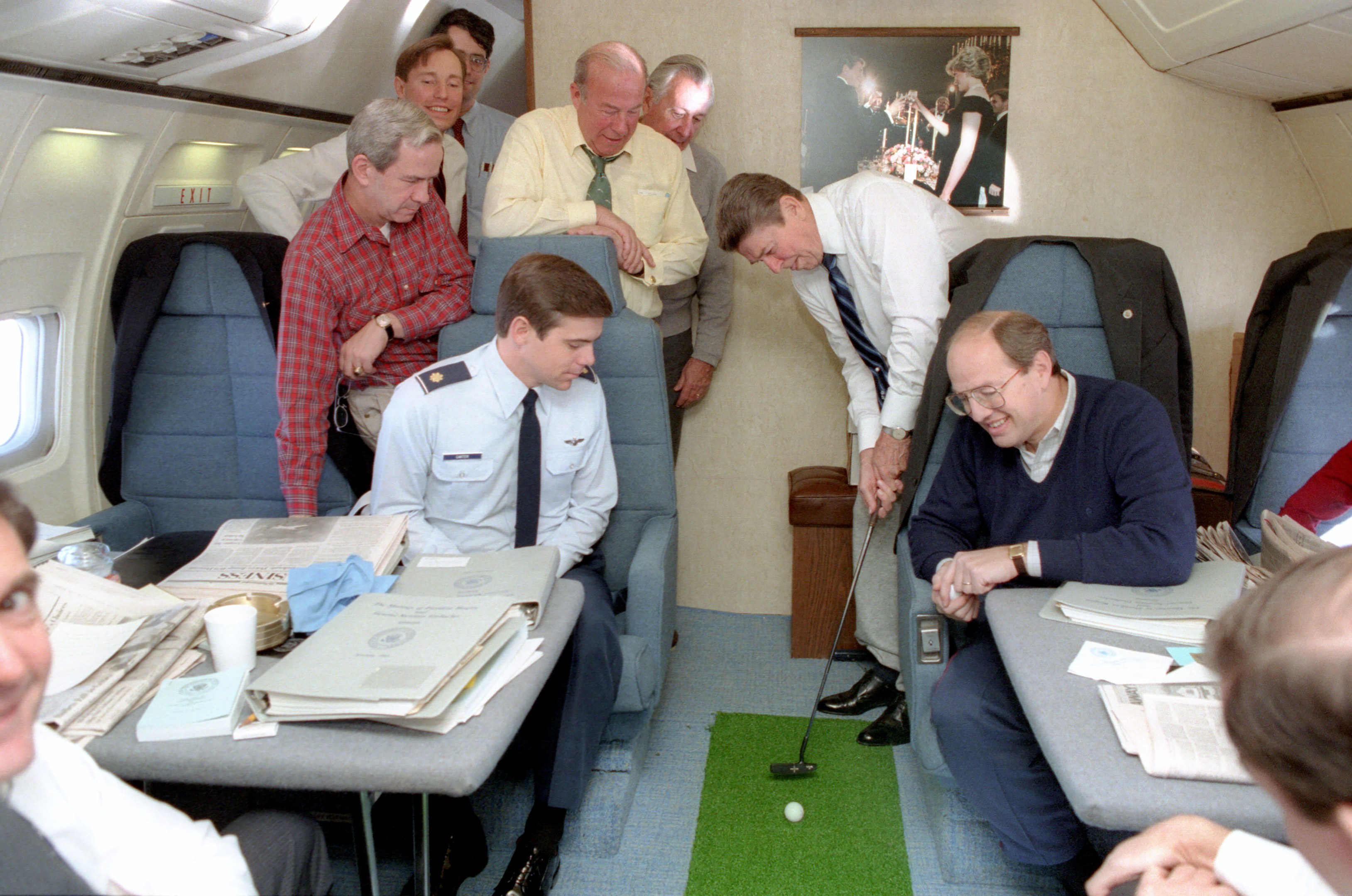 A group of men seated around a table in an airplane, with books, papers, cups, and other objects on the table, a photo frame on the wall, ceiling lights, and one man holding a golf club.