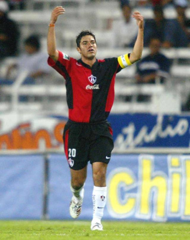 Ein Fussballspieler in rot-schwarzem Trikot rennt mit ausgebreiteten Armen auf einem Feld, umgeben von einer Zuschauermenge in den Rängen, mit einem "Chivas de Guadalajara"-Schild im Hintergrund.