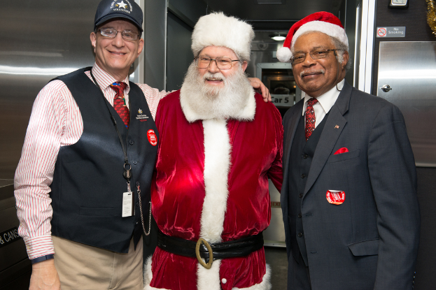 Eine Gruppe von Menschen mit Brillen, Mützen, Krawatten, Ausweisen und Abzeichen, darunter eine Person in einem Weihnachtsoutfit, die vor einer Stahlwand mit Lichtern, Schildern und anderen Gegenständen im Hintergrund steht.