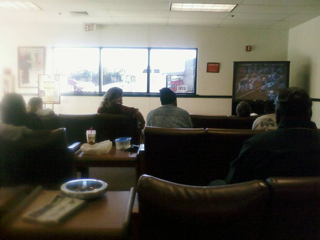 Ein Raum mit einer Decke, einem Fenster, einer Couch mit Menschen darauf, einem Tisch mit einer Tasse, einer Box, einer Abdeckung, einem Buch und einer Schüssel, einem Banner an der Wand, einem Mann mit einer Mütze und einem Ausgangsschild an der Decke.