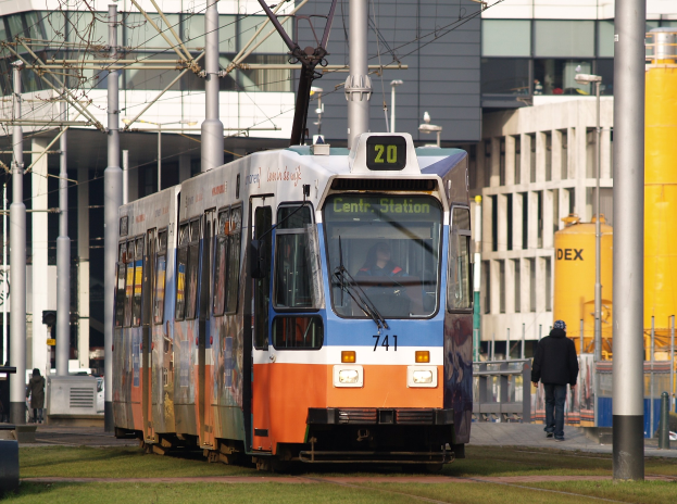 Blauer und orangefarbener Straßenbahnwagen auf einer Stadtstraße mit einem Passagier im Inneren, einer Person, die rechts auf dem Gehweg geht, und Gebäuden, Polen, Drähten und anderen Objekten im Hintergrund.