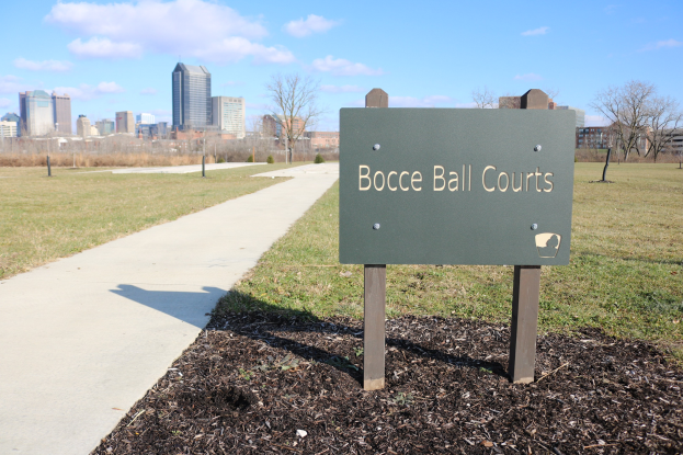 Ein Schild auf dem Boden in einem grasbewachsenen Bereich in der Nähe von Bäumen und Gebäuden trägt die Aufschrift "Bocce Ball Courts in Indianapolis, Indiana".