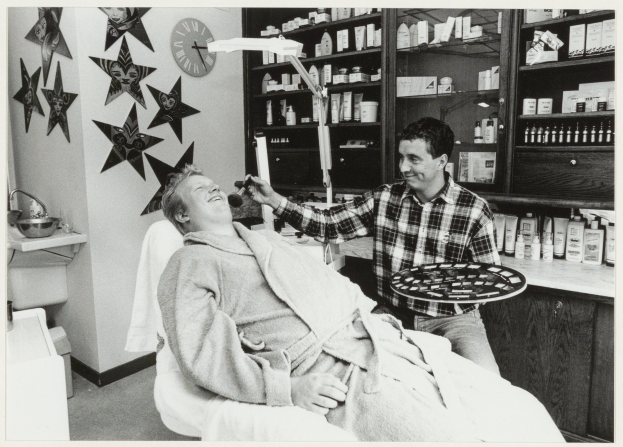 Ein Mann sitzt lachend in einem Zahnarztstuhl, während ein anderer Mann neben ihm steht und eine Platte hält, mit einem Waschbecken, Uhr, Sternen an der Wand und Regalen voller Flaschen im Hintergrund.
