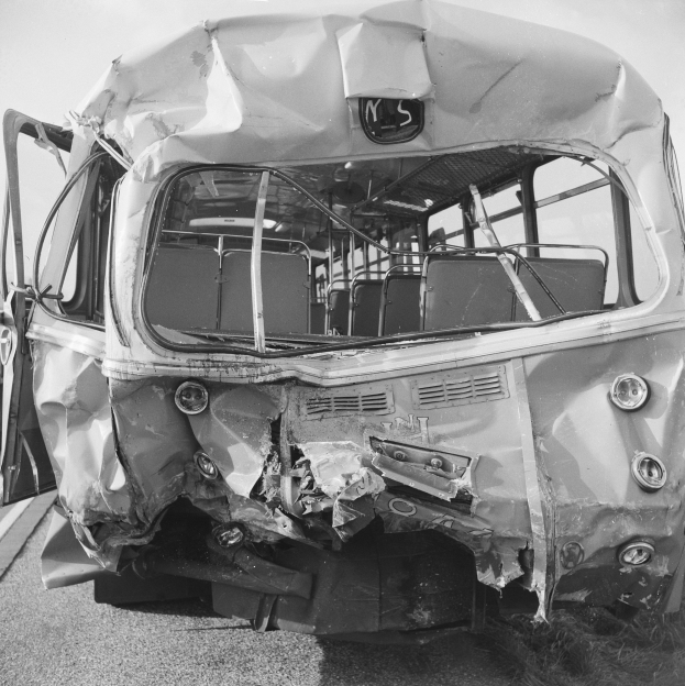 Ein Schwarz-Weiß-Foto eines schwer beschädigten Busses am Straßenrand, mit zerquetschter Front und zersplitterten Fenstern, unter einem klaren Himmel.