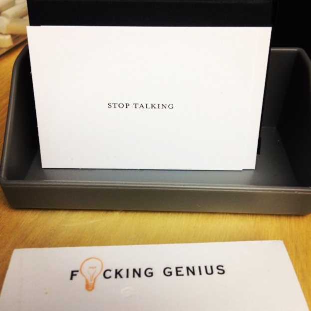 Ein Druckerbox mit einem Blatt Papier darauf, auf dem der Text 'Hör auf zu reden' steht, und einem weiteren Blatt Papier auf dem Tisch, auf dem der Text 'Verdammter Genius' steht.
