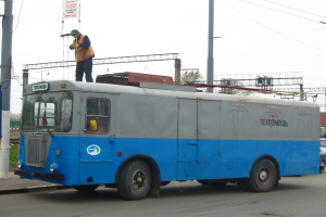 Ein Mann steht auf einem blauen und weißen Bus auf einer Straße mit anderen Fahrzeugen, Strommasten, einer Werbetafel, Bäumen, Gebäuden und einem klaren blauen Himmel im Hintergrund.