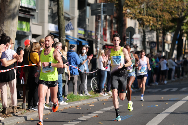 Eine Gruppe von Läufern bei einem Marathon auf einer Stadtstraße, mit Zuschauern auf der linken Seite, Bäumen, Gebäuden und einem klaren blauen Himmel im Hintergrund.