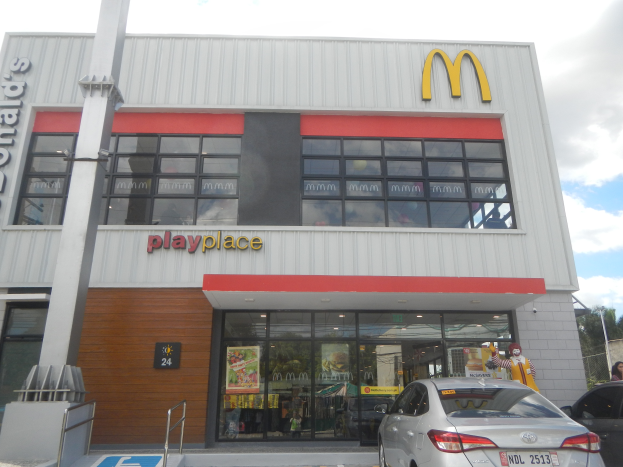 Ein McDonald's-Restaurant mit parkenden Autos davor, umgeben von Bäumen und Polen unter einem klaren blauen Himmel, mit Glaswänden, Fenstern und einem "McDonald's Playplace"-Schild, mit sichtbaren Postern und Gegenständen drinnen.