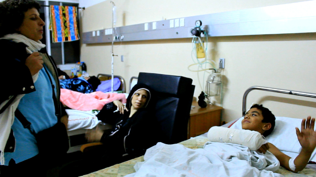 Ein Krankenhauszimmer mit mehreren Betten, jedes von einem Patienten belegt, mit KochsalzlÃ¶sungsflaschen auf StÃ¤nden zwischen den Betten und einer Frau mit einer Tasche in der NÃ¤he eines Patienten.