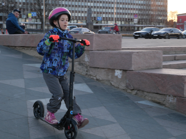 Ein junger Junge fährt mit einem Roller auf einem Gehweg, trägt einen Helm und Handschuhe und hat verschiedene urbane Elemente und einen klaren blauen Himmel im Hintergrund.