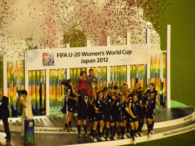 Gruppe von Frauenfußballspielerinnen in schwarzer Uniform, die auf einer Bühne feiern, umgeben von einer Menge mit Kameras, mit einem Hintergrundboard und dekorativen Gegenständen auf Gras.