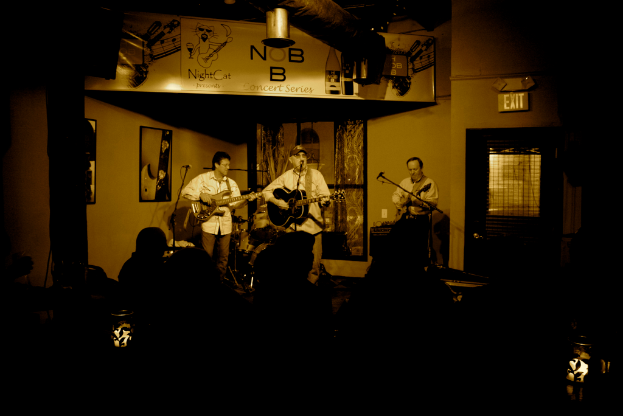 Eine Gruppe von Musikern in weißen Hemden, die auf einer Bühne mit einem Schlagzeug dahinter und einem Mikrofon davor Gitarren spielen, während das Publikum von vorne zusieht. Der Raum hat Gemälde an den Wänden und eine Tür mit einem Schild auf der rechten Seite.