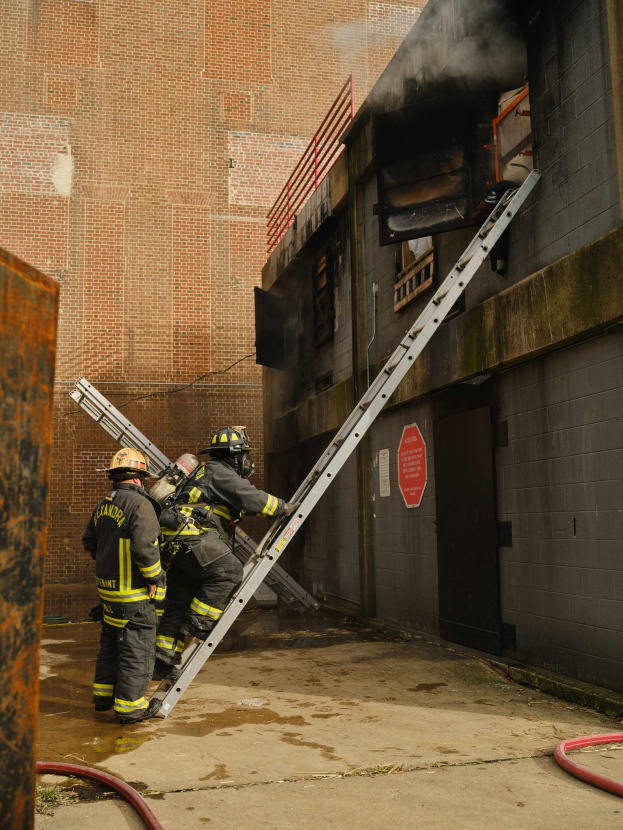 Feuerwehrleute in Helmen, die eine Leiter zu einem raucherfüllten Gebäude hinaufklettern, mit einem metallischen Objekt links und Rohren an der Basis.