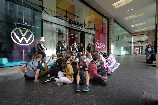 Eine Gruppe von Menschen sitzt vor einer Volkswagen-Vertretung mit Glaswänden und Logo auf dem Boden und hält eine Fahne mit Text in den Händen, unter Deckenlampen, scheinbar protestierend.
