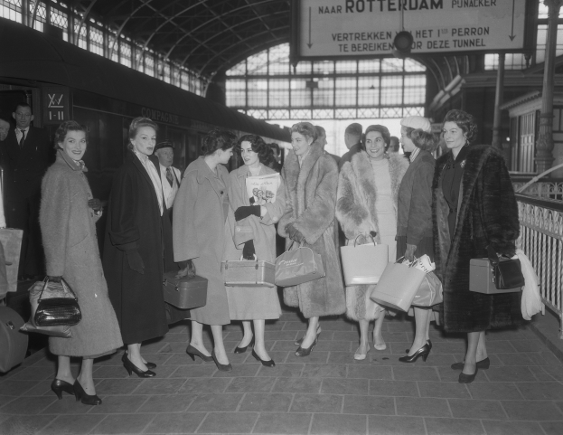Black and white image of women standing at a train station with a train on the left and railings and a board on the right, each holding a bag.
