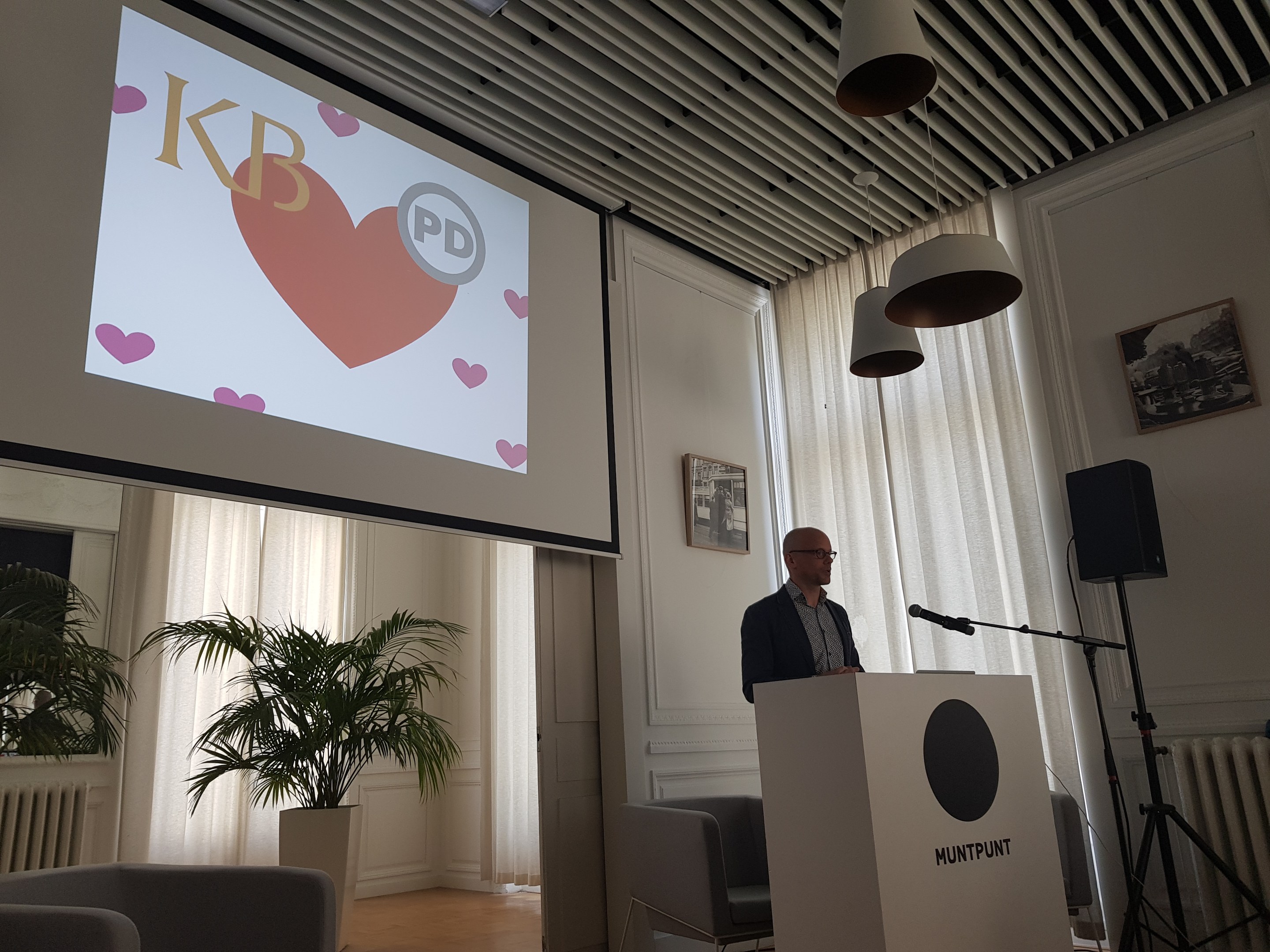 Ein Mann steht an einem Podium mit einem Mikrofon und spricht vor einem Publikum in einem Raum mit Stühlen, einer Pflanze, gerahmten Fotos an der Wand, Vorhängen, Deckenlampen und einem Projektionsschirm hinter ihm.