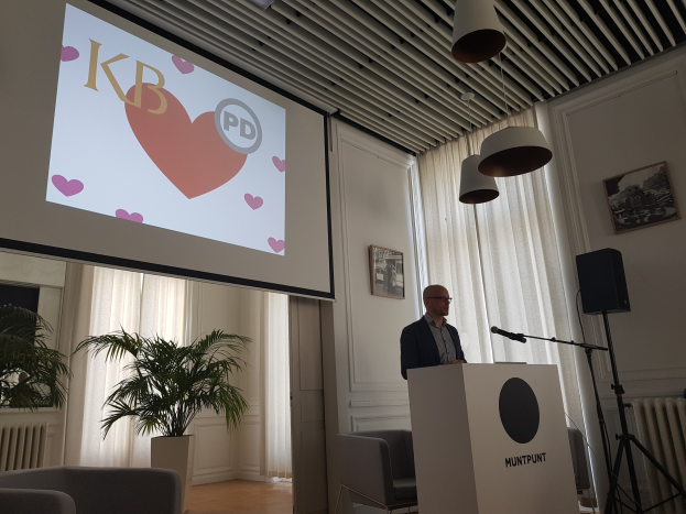 Ein Mann steht an einem Podium mit einem Mikrofon und spricht vor einem Publikum in einem Raum mit Stühlen, einer Pflanze, gerahmten Fotos an der Wand, Vorhängen, Deckenlampen und einem Projektionsschirm hinter ihm.