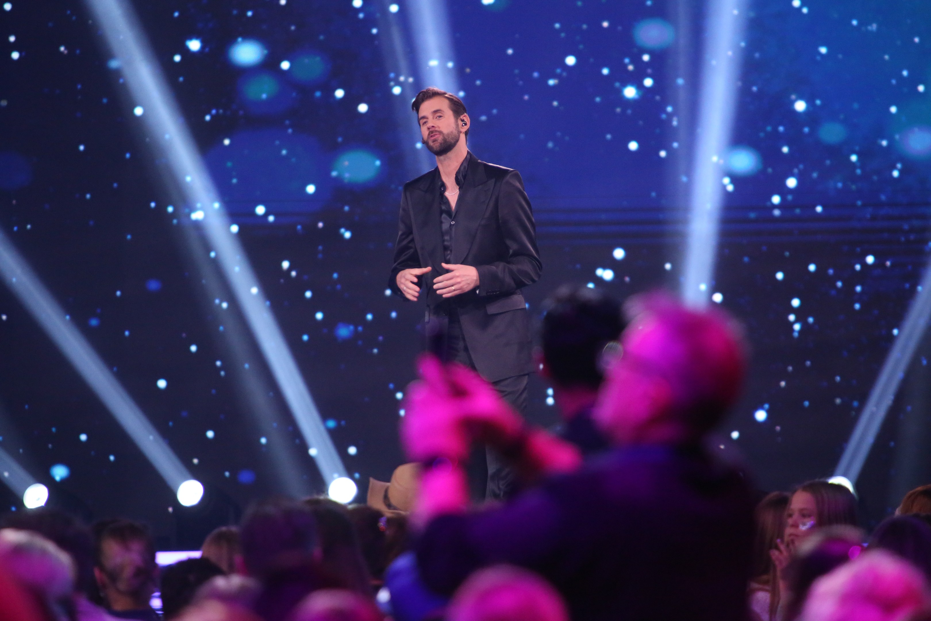 A man in a black suit stands on a brightly lit stage, holding a trophy, as the winner of *Dancing with the Stars* before a crowd.