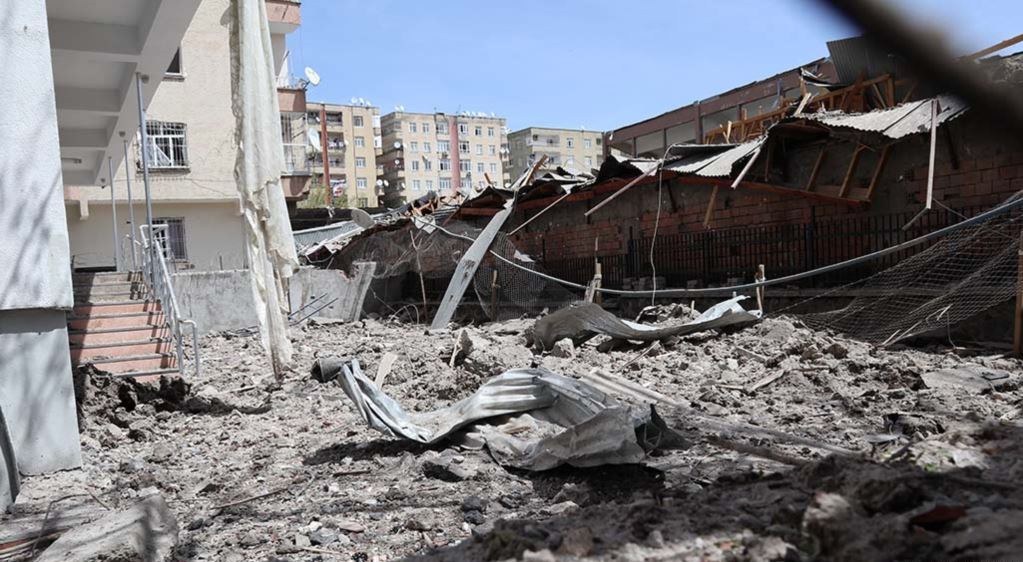 Die Nachwirkungen eines israelischen Luftangriffs in Gaza-Stadt zeigen ein eingestürztes Gebäude, das in der Mitte einer Straße zu Schutt reduziert wurde, mit freiliegenden Metallpfosten, einem Zaun, einer Treppe, benachbarten Gebäuden, Bäumen und einem bewölkten Himmel.