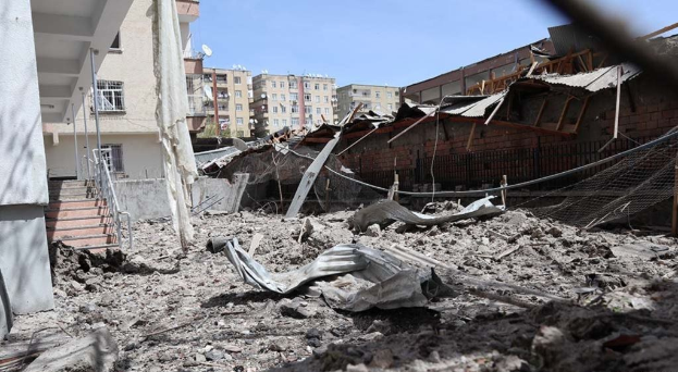Die Nachwirkungen eines israelischen Luftangriffs in Gaza-Stadt zeigen ein eingestürztes Gebäude, das in der Mitte einer Straße zu Schutt reduziert wurde, mit freiliegenden Metallpfosten, einem Zaun, einer Treppe, benachbarten Gebäuden, Bäumen und einem bewölkten Himmel.