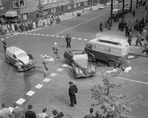 Eine Schwarz-Weiß-Szene eines Autounfalls an einer Straßenkreuzung mit mehreren Fahrzeugen, darunter ein Lieferwagen, einer Gruppe von Menschen drumherum und im Hintergrund Gebäude, Bäume, Pfähle und Texttafeln.