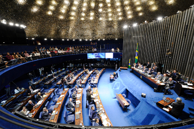 Eine Konferenzbühne mit sitzenden Personen an einem Tisch mit Papieren und Gegenständen, flankiert von Flaggen und einer Skulptur im Hintergrund, mit Sitzplätzen im Vordergrund und Stehplätzen hinter einem Geländer mit Bildschirmen und Deckenbeleuchtung.
