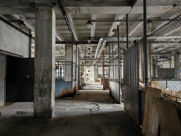 Leerer Raum mit großen Fenstern, Säulen, einer Tür, Deckenleuchten, Metallstangen, einem Maschendrahtzaun, verstreuten Gegenständen auf dem Boden und einer Wand, die Zeichen von Verlassenschaft und Vernachlässigung trägt.