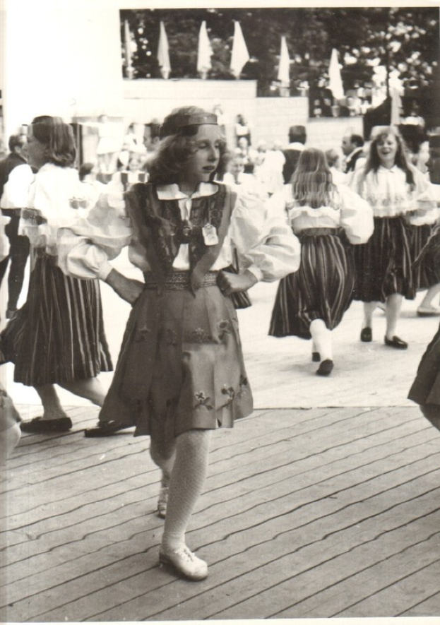 Eine Gruppe von Frauen in traditioneller bayrischer Tracht tanzt auf einem Holzboden, mit einer Wand, Bäumen und anderen Gegenständen im Hintergrund. Text ist unten im Bild sichtbar.