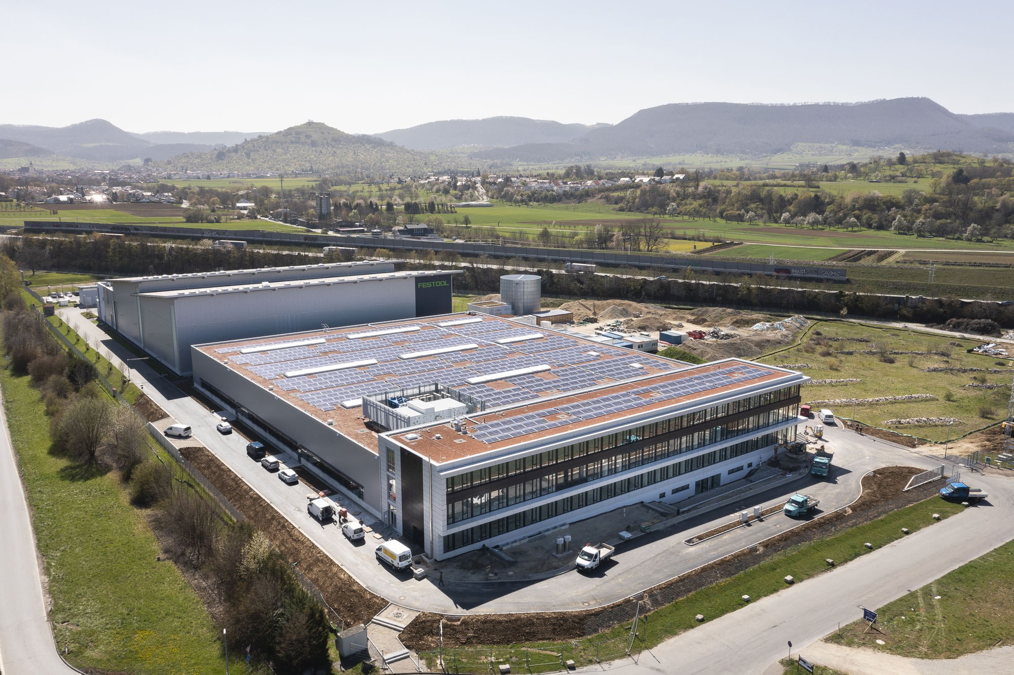Aerial view of a large building with solar panels on its roof, surrounded by vehicles, trees, grass, and poles, with hills and a clear blue sky in the background.