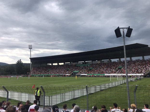 Große Zuschauermenge bei einem Fußballspiel in einem Stadion mit bewölktem Himmel, beleuchtetem Feld und umliegenden Bäumen und Hügeln.