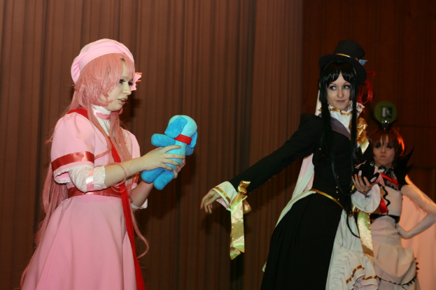 Drei Frauen führen ein Drama auf, wobei eine in einem rosafarbenen Kleid ein blaues Teddybär hält, eine andere in einem schwarzen Kleid nach links schaut und ein brauner Vorhang im Hintergrund zu sehen ist.