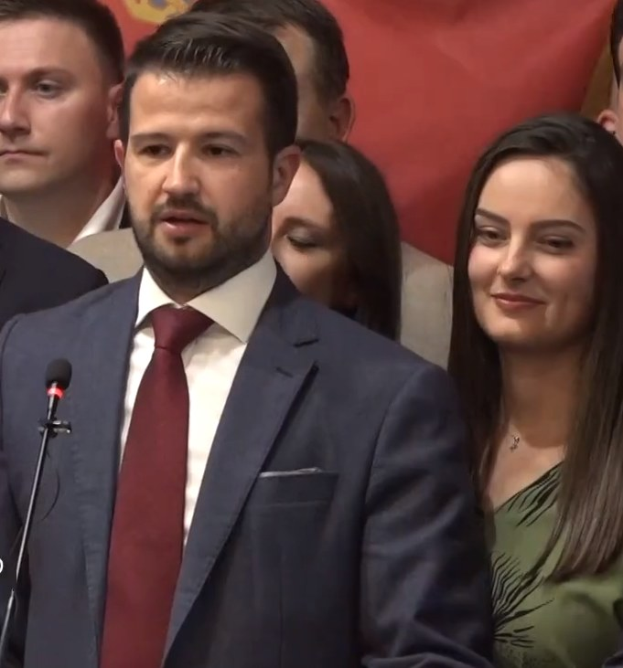 A suited man speaks at a podium with a microphone, addressing a crowd behind a banner, likely discussing a government social media ban.