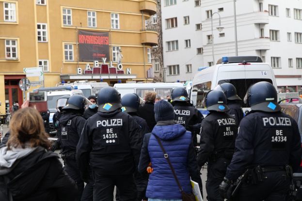 Polizeibeamte in Uniform vor einer Menge bei einer Demonstration in Berlin, mit Fahrzeugen, Gebäuden und einem Kameramann im Hintergrund.