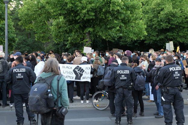 Große Gruppe von Menschen an einer Straße mit Schildern und Mützen und Taschen, mit einem Fahrrad im Vordergrund und Bäumen und einem Pfahl im Hintergrund während einer Black-Lives-Matter-Demonstration in Berlin.